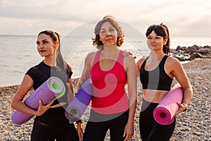 Portrait of group of Caucasian pretty adult women holds sports mats on pebble beach. Sea and wild coast in background