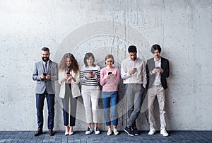Portrait of group of entrepreneurs standing against concrete wall indoors in office, using smartphone.