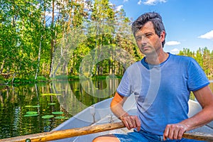 Portrait of handsome adult man holding oars in hands and floating on boat on northern lake. Happy vacationer sitting in boat