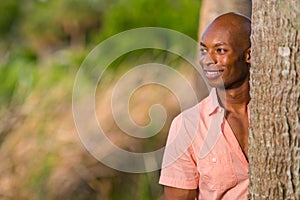 Portrait handsome African American man posing from behind a tree in the park. Man is smiling and glancing off camera