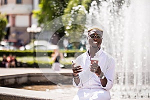 Portrait of handsome afro american young man listening music from phone drinking coffee to go on summer street in front of fountai