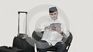 Portrait of handsome asian muslim man with head cap sitting on couch and using tab