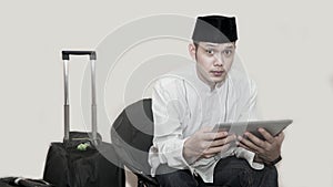 Portrait of handsome asian muslim man with head cap sitting on couch and using tab