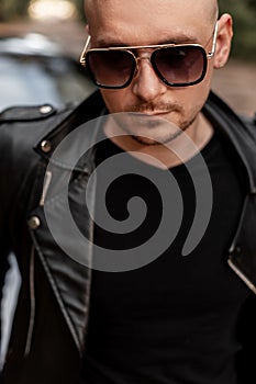 Portrait handsome bald man with beard in sunglasses in fashion leather jacket near modern black car outdoors. Cool guy rest next