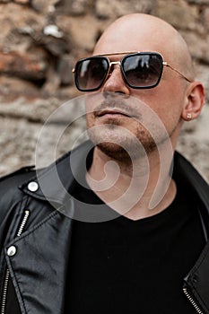 Portrait handsome bald man in stylish sunglasses in casual trendy black leather jacket near brick wall on street. Cool fashion guy
