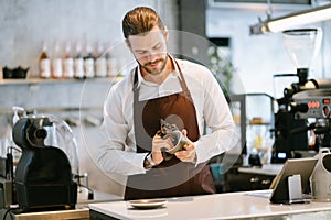 Portrait of handsome barista standing in behind the counter bar and smiling while wiping ceramic cups in a coffee shop. Clean