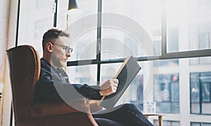 Portrait handsome bearded businessman wearing glasses,black shirt.Man sitting in vintage chair modern loft studio, reading book an