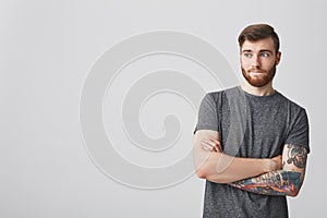 Portrait of handsome bearded hipster guy with dark hair and tattoo sleeve looking aside with crossed hands and cynical