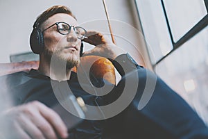 Portrait handsome bearded man glasses,headphones listening to music modern loft studio.Man sitting in vintage chair,holding smartp