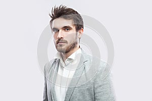Portrait of handsome bearded serious man with brown hairs and beard in white shirt and casual grey suit standing and looking at