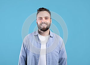 Portrait of handsome bearded young man in casual outfit smiling and looking at camera over blue studio background