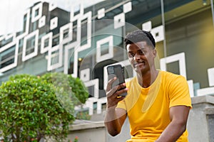 Portrait of handsome black African man wearing yellow t-shirt outdoors in city while using mobile phone