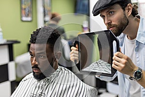 Portrait of handsome black man with comb in hair looking in the mirror at his new haircut. Barber hairdresser showing client his