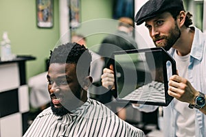 Portrait of handsome black man with comb in hair looking in the mirror at his new haircut. Barber hairdresser showing client his