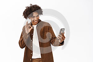 Portrait of handsome boy video chat, talking on mobile phone application, standing over white background, showing okay