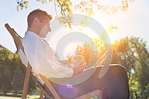 Portrait of handsome businessman drinking coffee and reading newspaper while sitting on folding chair in park
