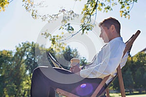 Portrait of handsome businessman drinking coffee and reading newspaper while sitting on folding chair in park