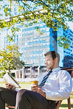 Portrait of handsome businessman drinking coffee and reading newspaper while sitting on folding chair in park