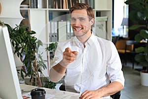 Portrait of a handsome businessman in white shirt pointing finger at camera while sitting on the table at office