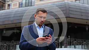 Portrait of handsome Caucasian male walking in city while using smartphone. Young successful gentleman texting message