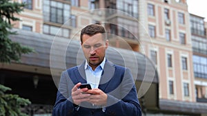 Portrait of handsome Caucasian male walking in city while using smartphone. Young successful gentleman texting message