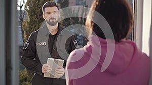 Portrait of handsome caucasian police officer standing with tablet at entrance door and talking to brunette woman