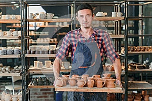 Portrait of a handsome caucasian pottery worker standing with tray full of ceramic pots at the pottery manufacturing.