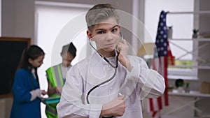 Portrait of handsome Caucasian teenage boy in medical gown using stethoscope looking at camera smiling. Happy satisfied