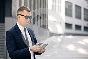 Portrait of handsome caucasian young businessman in sunglasses counting money while standing outdoors. Joyful male with