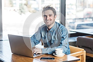 Portrait of handsome cheerful bearded young businessman in blue jeans shirt are sitting in cafe and typing message on laptop with