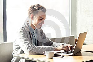 Portrait of handsome cheerful bearded young businessman in grey blazer are sitting in cafe and typing message with toothy smile