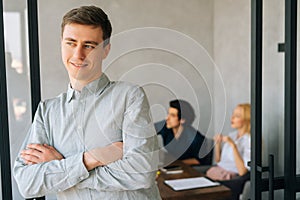 Portrait of handsome confident young business man in casual clothes standing with arms crossed in office smiling looking