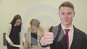 Portrait of a handsome confident young man smiling looking in camera while his female colleagues working with papers in