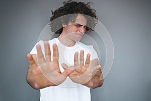 Portrait of handsome curly hair male shows refusal and stop gesture, has angry and unhappy expression, wearing white t-shirt,