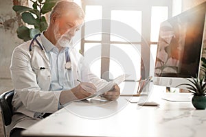 Portrait of handsome doctor sitting at desk, taking notes or fills in the client& x27;s medical card or prescribes medication