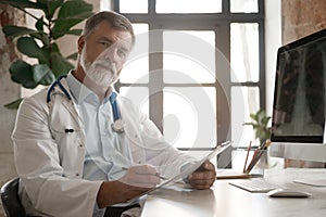 Portrait of handsome doctor sitting at desk, taking notes or fills in the client's medical card or prescribes medication