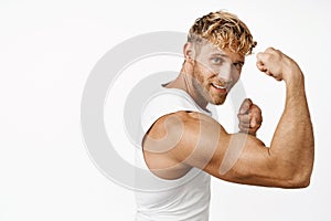 Portrait of handsome fitness athlete flexing biceps, showing strong muscle arm and smiling, workout in gym, standing