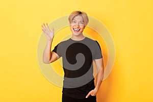 Portrait of handsome friendly asian guy in black outfit, waving hand to say hello and smiling, greeting someone