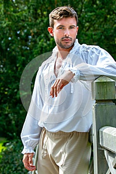 Portrait of handsome gentleman dressed in vintage costume standing on bridge of stately home with trees in background