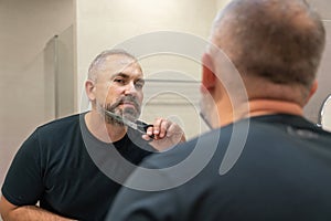 Portrait of handsome gray-haired man in his 40s trimming his beard himself standing in bathroom