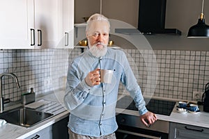 Portrait of handsome gray-haired mature adult male drinking coffee and looking at camera in morning at home. Thinking