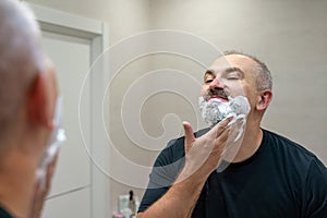 Portrait of handsome gray-haired middle-aged man applying shaving foam to trim his beard