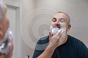 Portrait of handsome gray-haired middle-aged man applying shaving foam to trim his beard