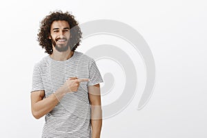 Portrait of handsome guy with charming smile and curly hair in casual clothes, pointing right and smiling with assured