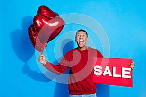 Portrait of handsome guy hold sale banner heart figure balloons look up empty space isolated on blue color background