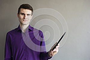 Portrait of a handsome guy looking away at the tablet. In a purple shirt. Brunette with green eyes. On a monophonic gray backgroun