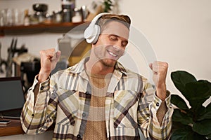 Portrait of handsome happy guy, freelancer working and listening to music in wireless headphones, dancing on his chair