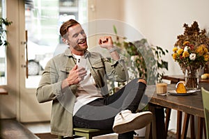 Portrait of handsome happy man in cafe, listens to music in wireless earphones, holding smartphone, connects to public