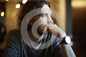 Portrait of handsome hipster guy with long hair and beard sitting in sunny cafe, resting near window. Romantic man looks lonely.