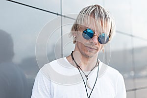 Portrait of handsome hipster young man in stylish white t-shirt with trendy hairstyle in vintage sunglasses near a gray modern